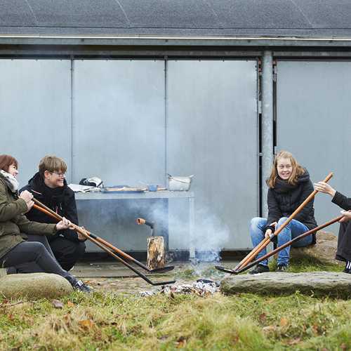 Fem elever sidder rundt om et bål på en græsplæne. Eleverne bager pandekager med hver sin bålpande på en lang pind. Eleverne er iført jakker, og det ser ud til at være efterår.