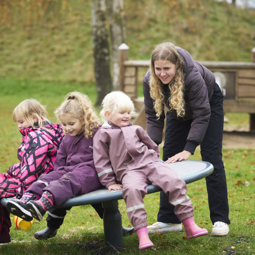 En pædagogisk assistent hjælper tre børn, som sidder tæt sammen på et vippebræt på legepladsen. Alle smiler og er iført regnsæt. 