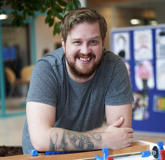 Mandlig skoleelev iført grøn T-shirt læner sig op ad et bordfodboldbord. Han smiler med ansigtet rettet mod kameraet.